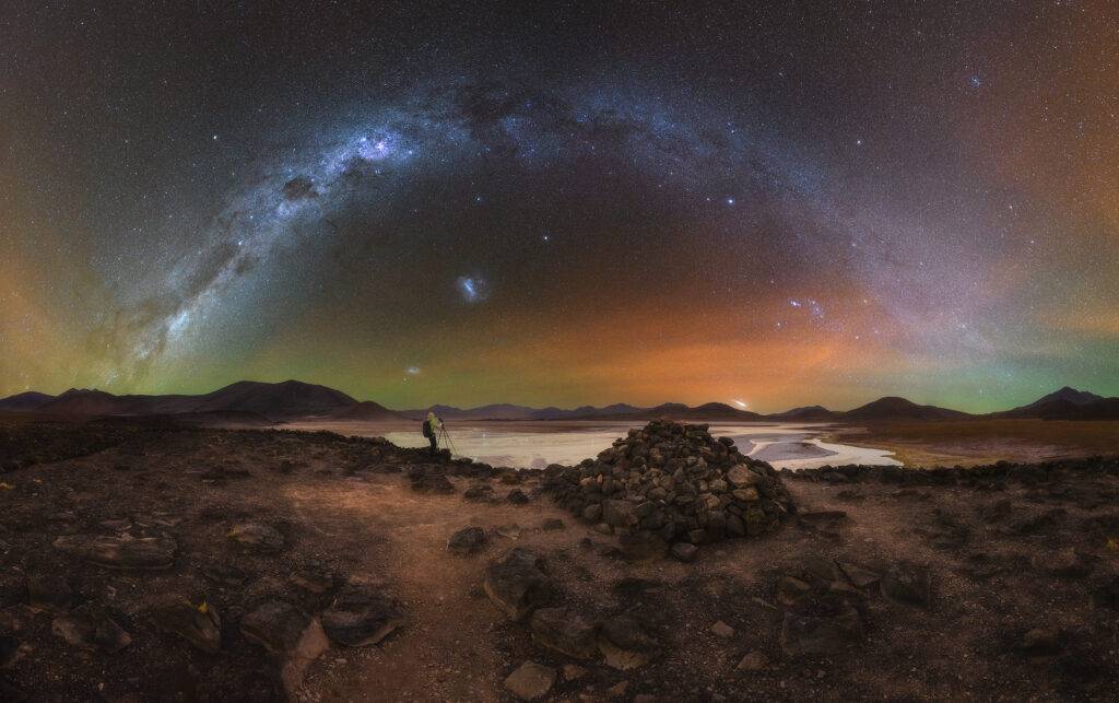 Fotógrafo con trípode bajo el arco completo de la Vía Láctea en el Mirador de Aguas Calientes, con salar y volcanes al fondo, San Pedro de Atacama