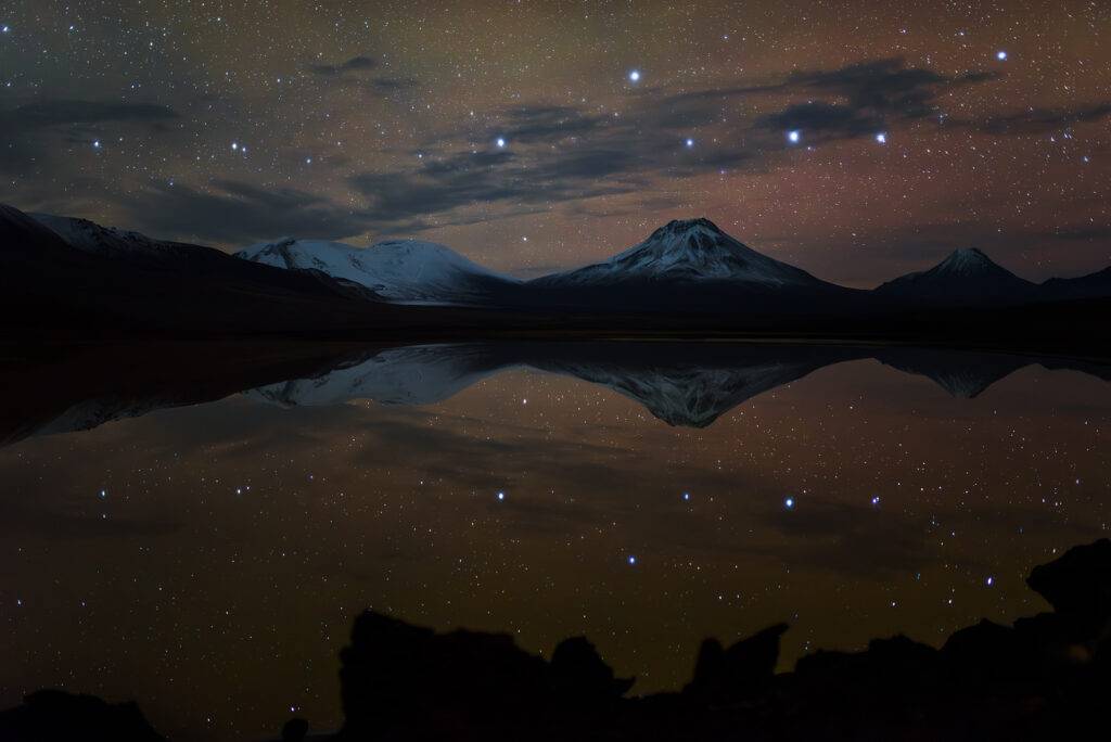 Reflejo nocturno de volcanes nevados en la Laguna Lejía, Atacama, con estrellas reflejadas en el agua en calma