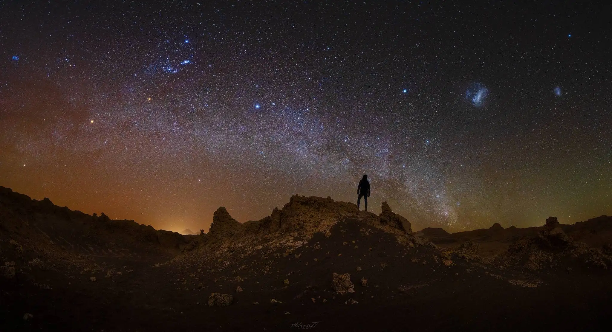 Silueta de fotógrafo sobre formaciones rocosas del desierto de Atacama bajo el cielo de verano, con la constelación de Orión, el Brazo de Orión y las Nubes de Magallanes visibles, noviembre-diciembre