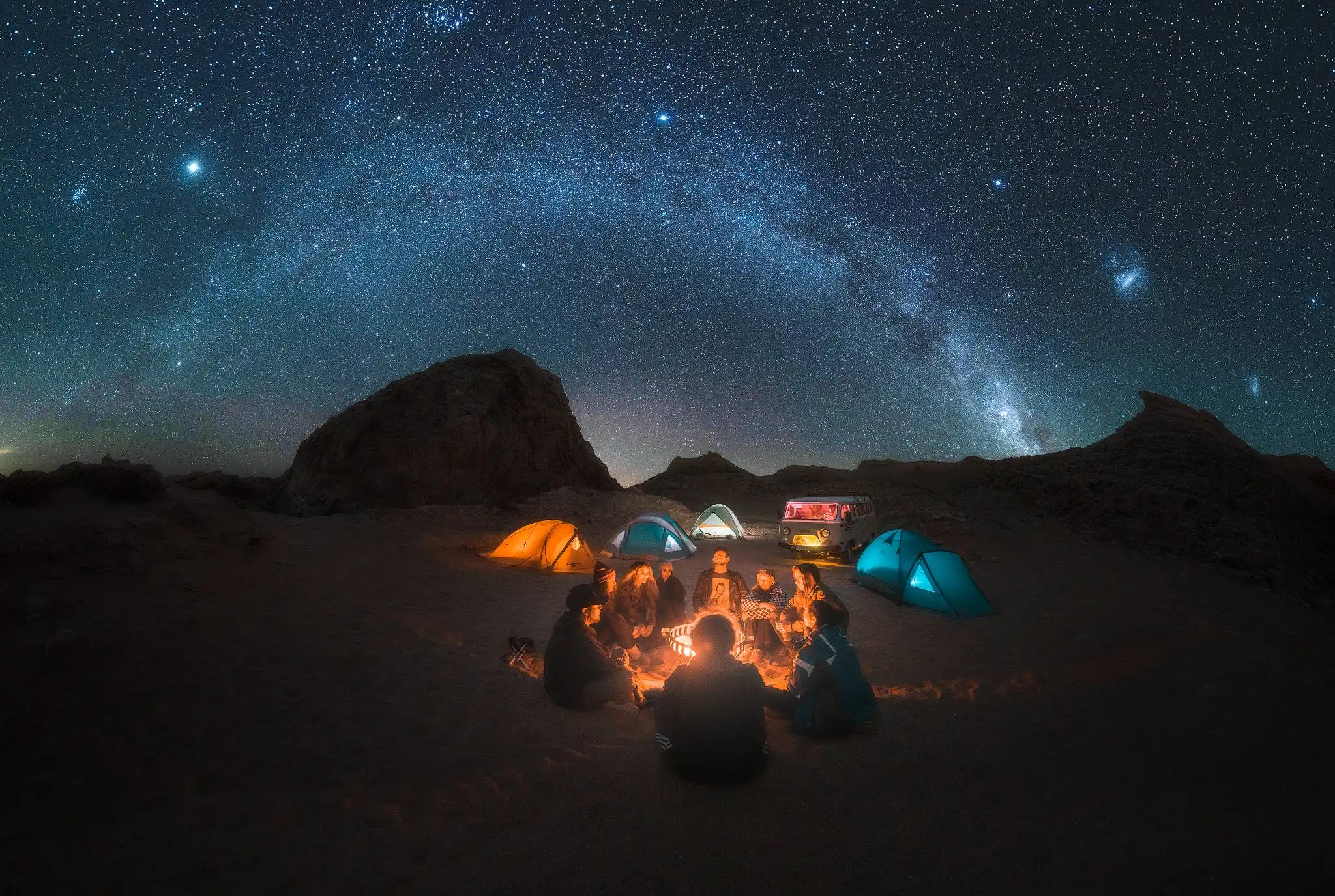 Grupo de fotógrafos reunidos alrededor de una fogata bajo el cielo de verano en Atacama, con el Brazo de Orión y las Nubes de Magallanes visibles sobre el desierto, San Pedro de Atacama